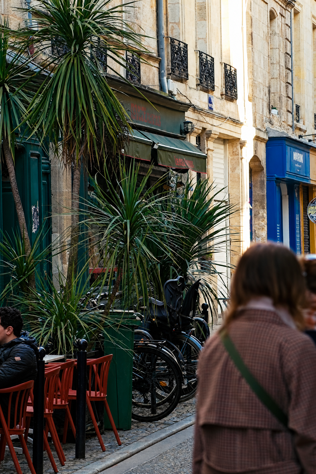 Sunlit Bordeaux street with cafe tables, palm trees and people walking past old stone buildings