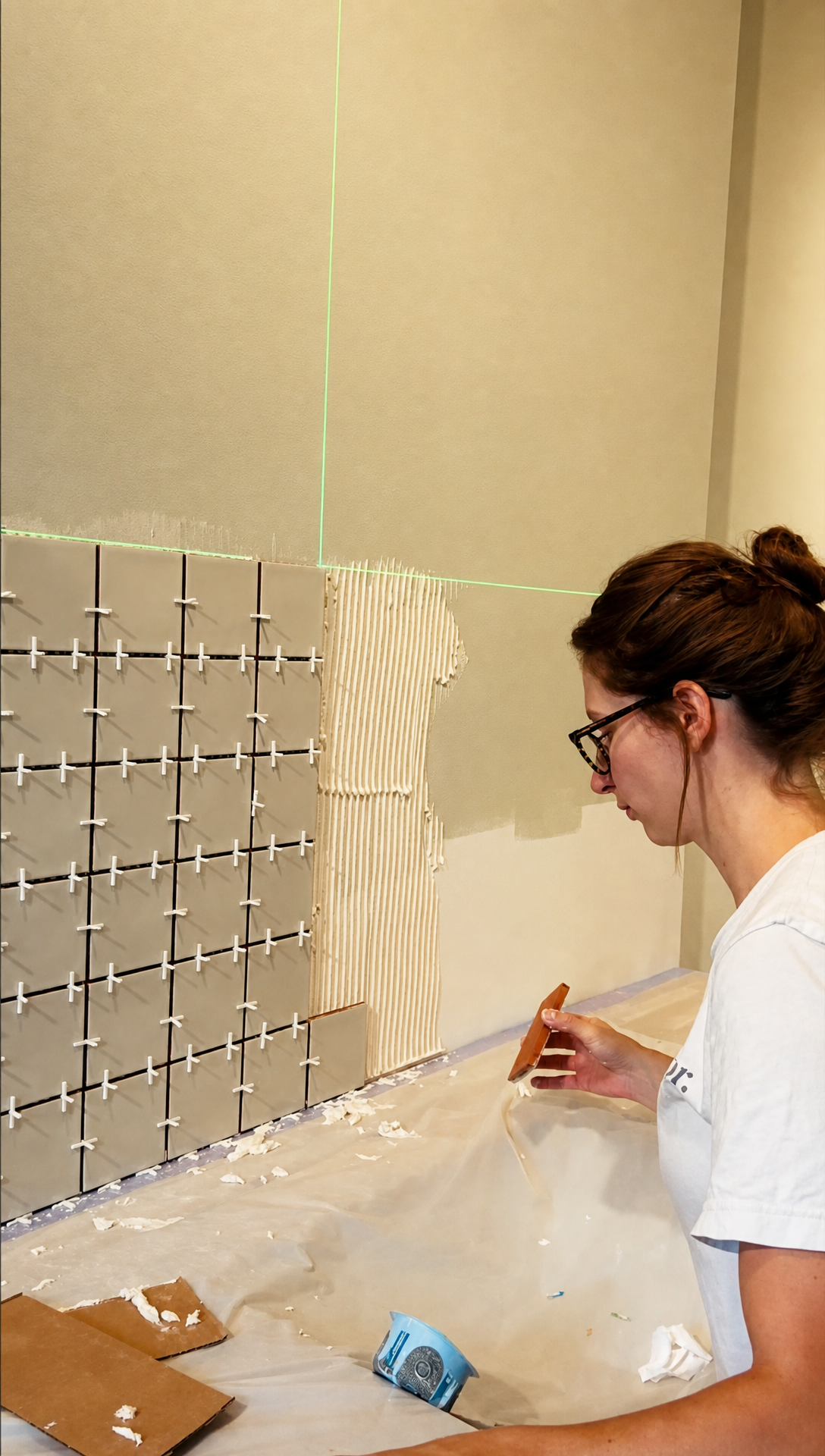 Romy laying wall tiles with spacers, guided by a green laser level line