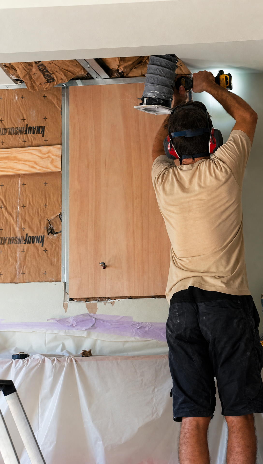 Hugo screwing a plywood reinforcement panel into the opened wall with flexible ducting hanging from the ceiling above
