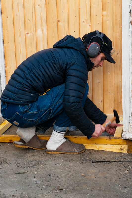 Hugo crouched in the greenhouse doorway with ear defenders on, hammering the base timber below the fresh wooden boarding