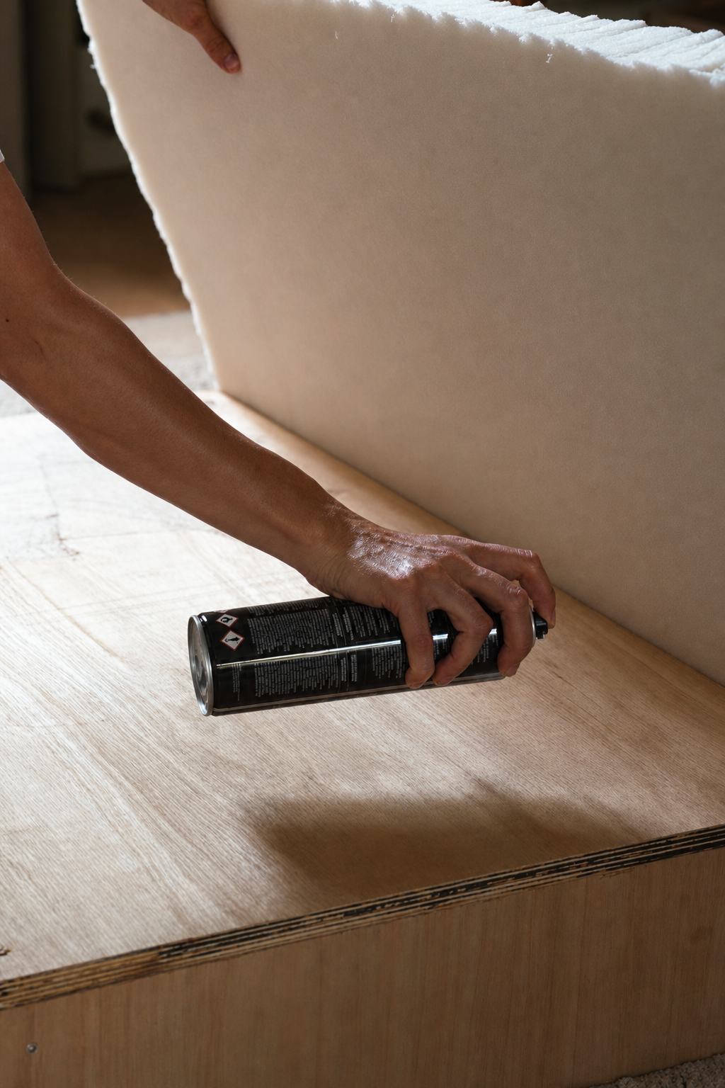 A hand spraying glue onto the wooden footstool box with the foam slab held behind
