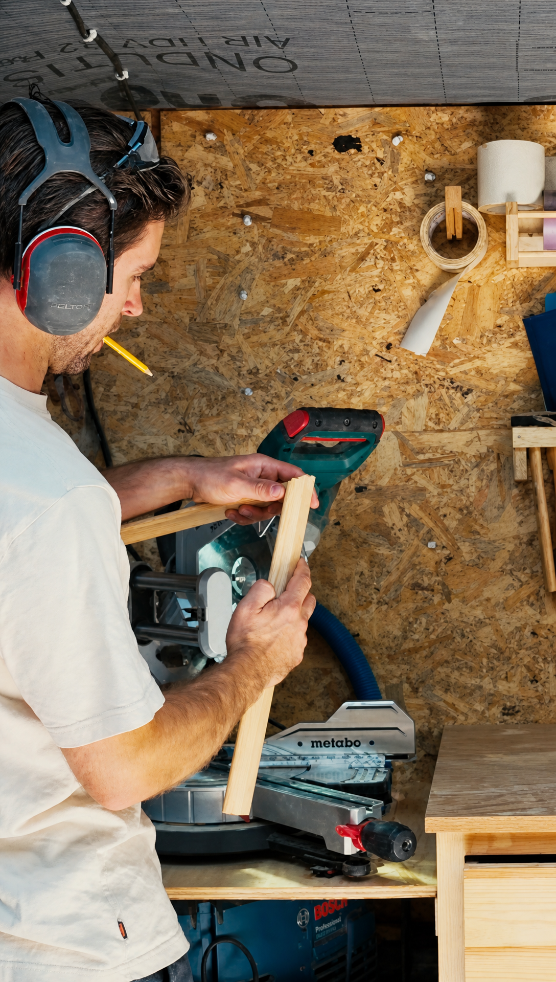Hugo in the workshop wearing ear defenders, pencil in mouth, checking a length of moulding beside the mitre saw