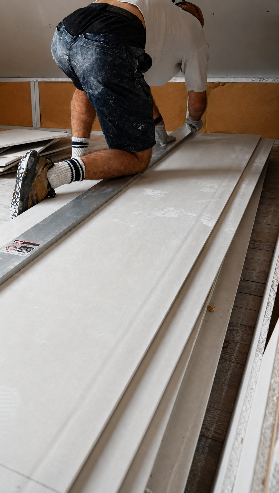 Hugo kneeling on a stack of plasterboard sheets, holding a long metal straight edge in place to cut along