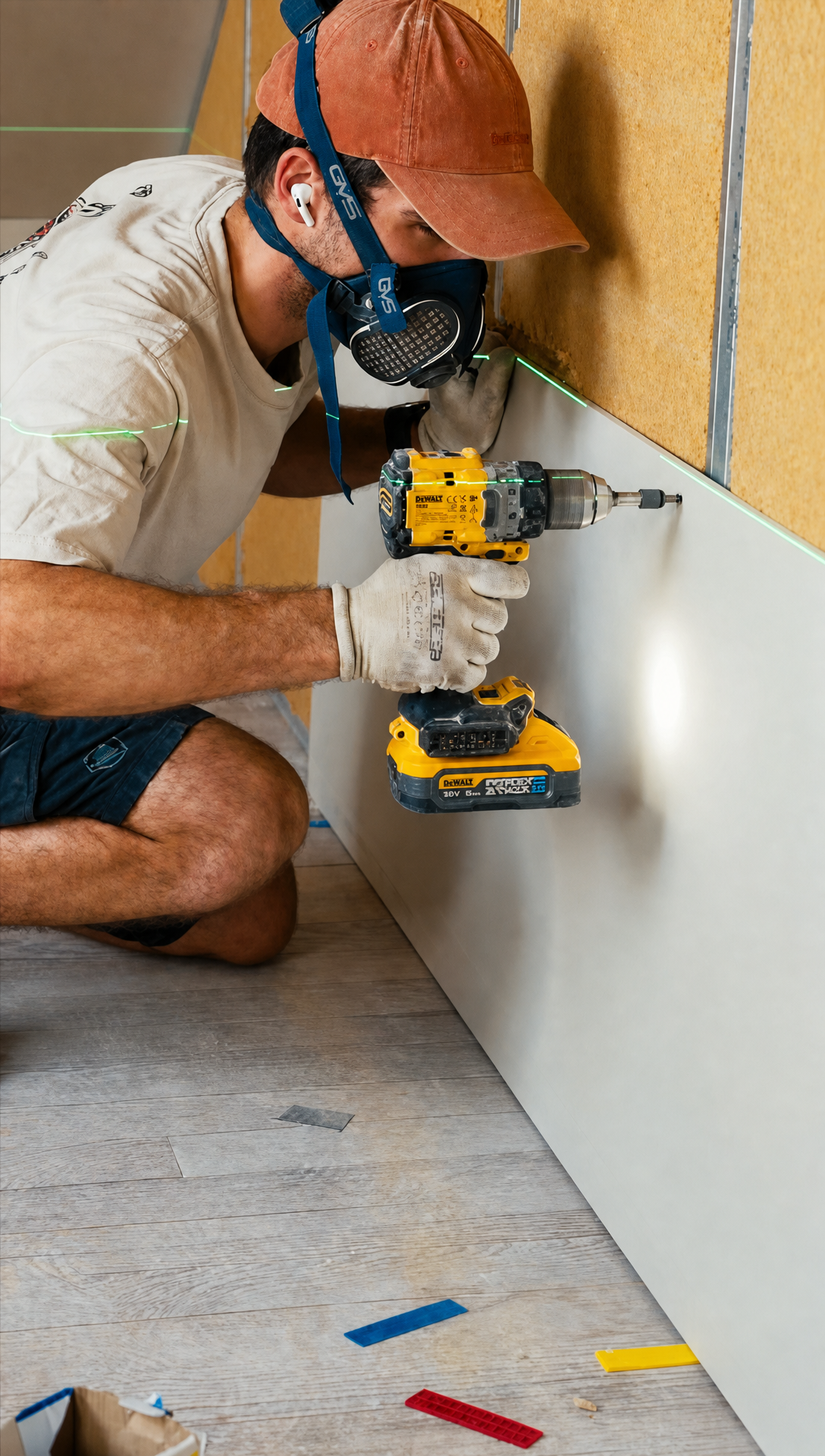 Hugo in a cap and dust mask crouching to screw the bottom edge of a plasterboard sheet to the wall, a green laser line running across it