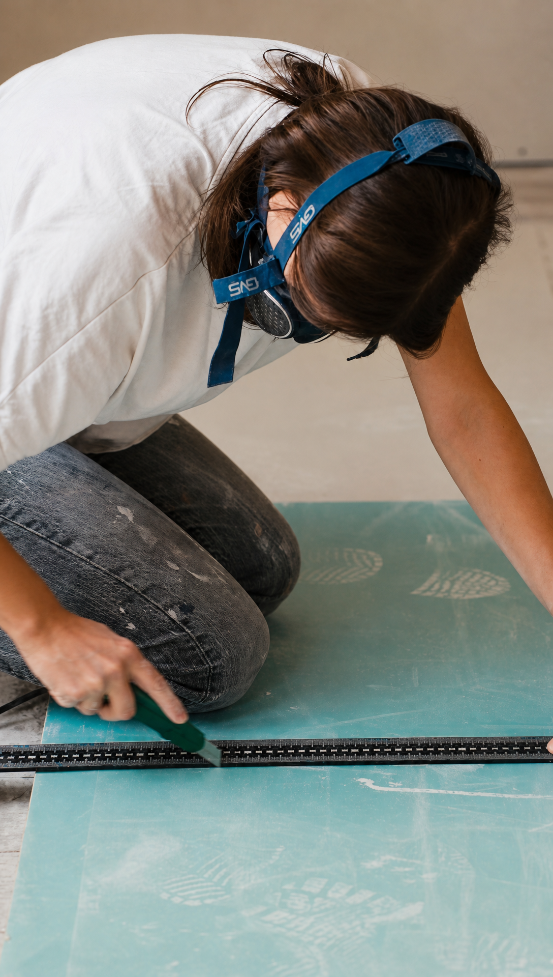 Romy in a dust mask kneeling on a sheet of green plasterboard, scoring it with a knife along a metal rail