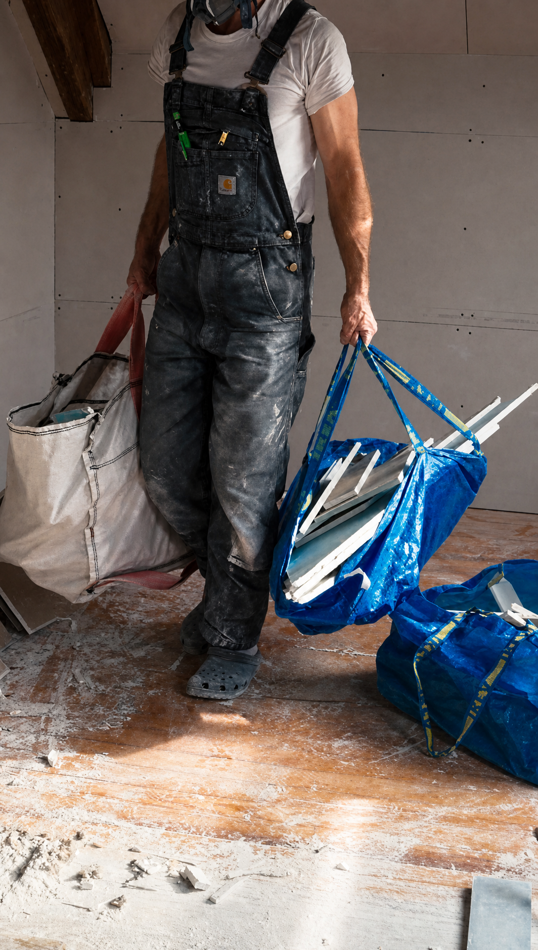 Hugo in dusty dungarees carrying two large bags of plasterboard offcuts across the attic floor