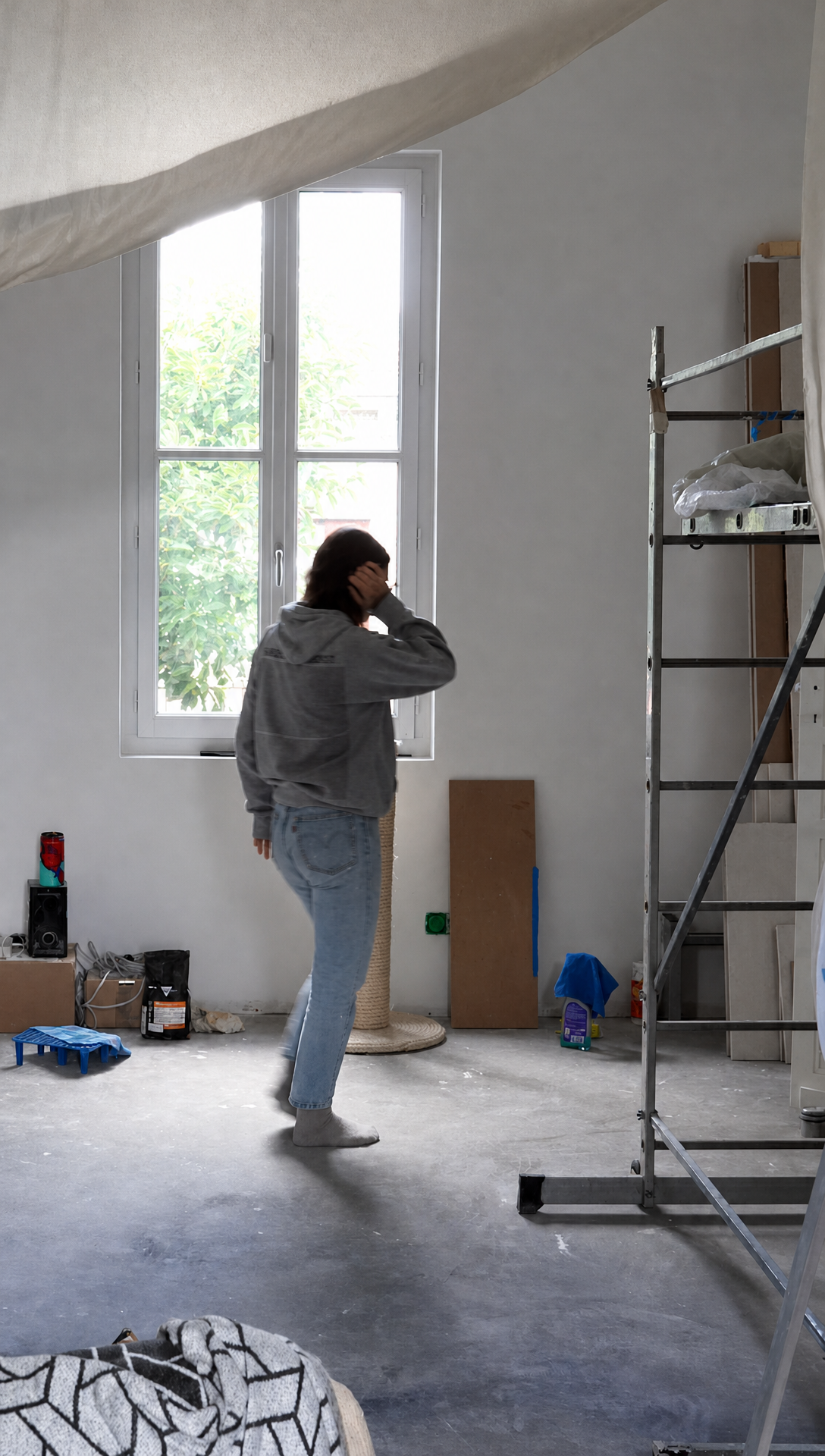 The empty living room before renovation, with bare walls, a concrete floor and a scaffold tower by the window