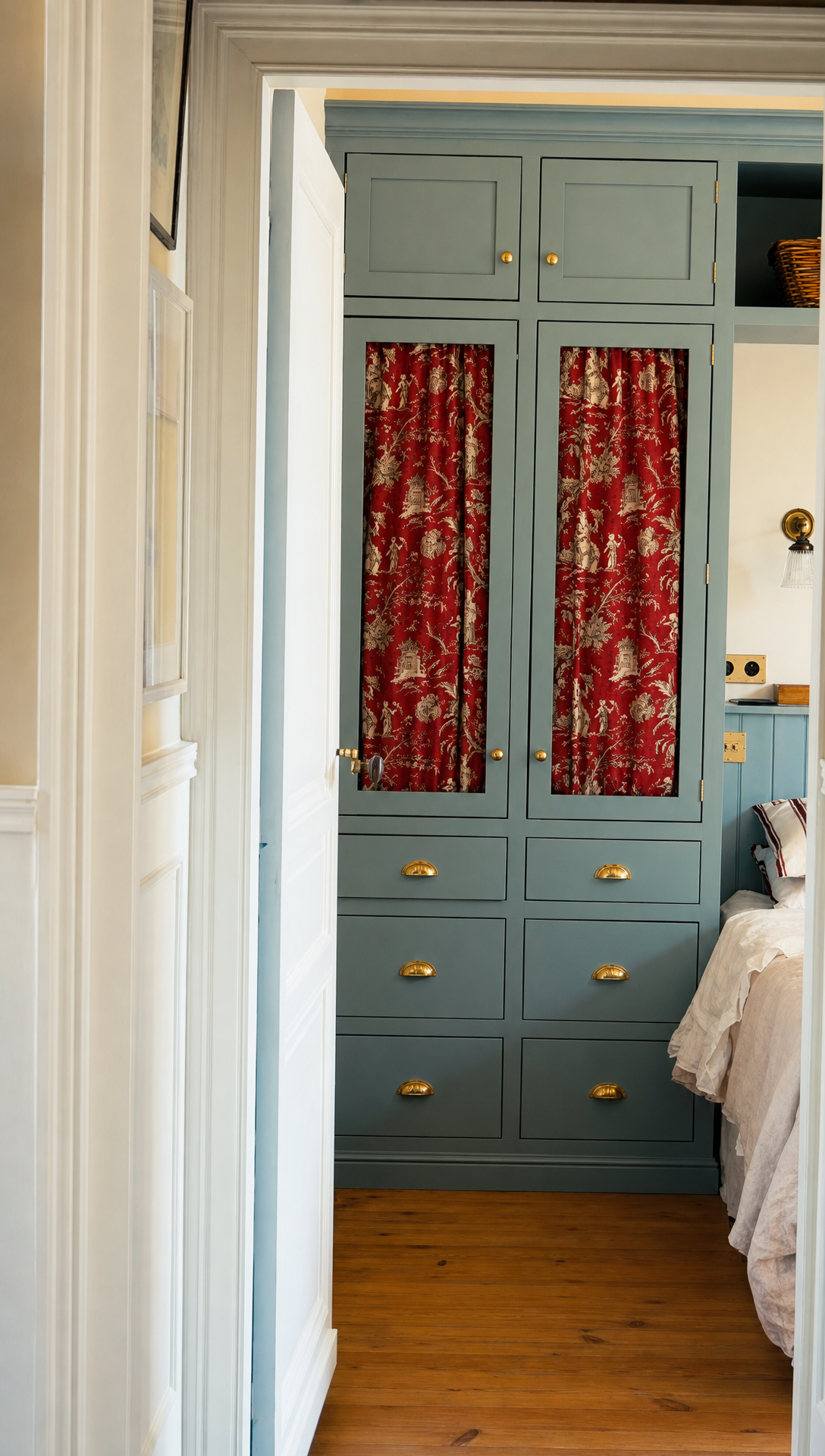 View through the bedroom door to the built-in wardrobes with red toile curtain panels and brass handles, and the bed between them